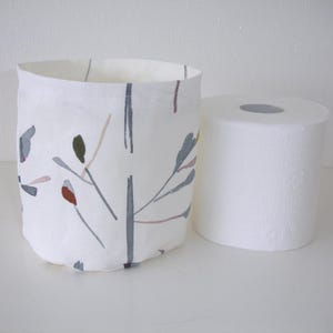 May include: A cylindrical fabric storage basket featuring a white base with a printed pattern of thin grey branches, muted green leaves, and small reddish-brown accents, positioned next to a standard roll of white toilet paper.
