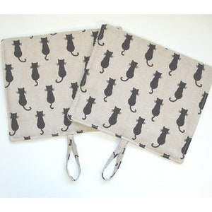 May include: Two square beige pot holders with a repeating pattern of black cat silhouettes. Each pot holder has a loop for hanging. The pot holders are made of a natural-looking fabric.