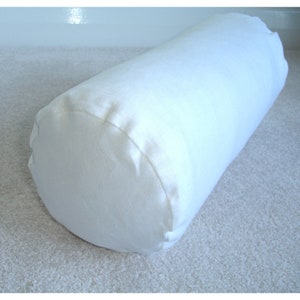 May include: A white cylindrical pillow, made of a soft, textured fabric. The pillow is lying on a white carpet.
