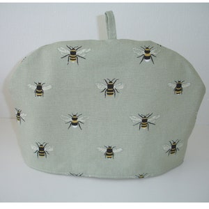 May include: A green tea cozy with a bee pattern. The cozy is made of fabric and has a handle on top.