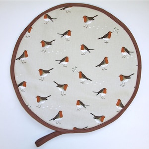 May include: Circular beige fabric item with a brown trim and handle. The fabric features illustrations of robins with red breasts and brown wings. Some robins hold sprigs. Likely a kitchen accessory, perhaps a pot stand or trivet.