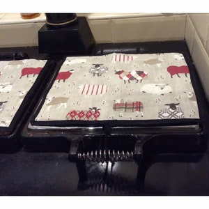 May include: Two stovetop protectors with a sheep pattern. The protectors are made of a beige fabric with red, white and black sheep wearing sweaters. The protectors are on a black stovetop.