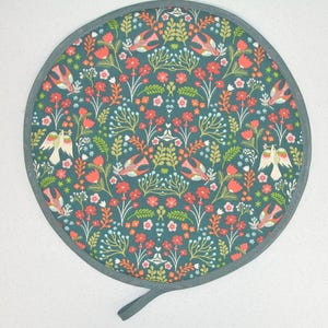 May include: Circular teal pot holder with a floral and bird design. It features red flowers, green foliage, and stylized birds. The edge is trimmed with grey binding and a hanging loop.