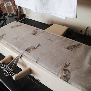 May include: A beige stovetop cover with a pattern of brown rabbits and white dots. The cover is made of fabric and has a white border.