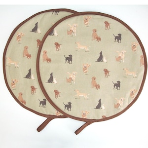 May include: Two circular placemats with a brown trim and handle. The beige fabric is patterned with illustrations of dogs in various poses and colours, including brown, black, and tan.