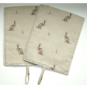 May include: Two beige linen pot holders with a brown rabbit pattern and white polka dots. Each pot holder has a loop handle.