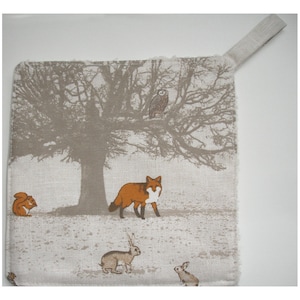 May include: A beige fabric pot holder with a woodland scene featuring a fox, squirrel, owl, and rabbits. The background includes a tree with bare branches.