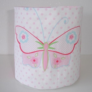 May include: A cylindrical fabric lampshade featuring a white background with a pattern of small pink polka dots. A large butterfly graphic is centered on the front, colored in shades of pink, light blue, and green with floral wing details.