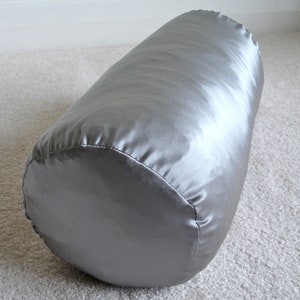 May include: A cylindrical bolster pillow in a silvery-gray satin fabric. The pillow is smooth and has a slight sheen, with visible stitching along the rounded ends. It rests on a light-colored carpet.