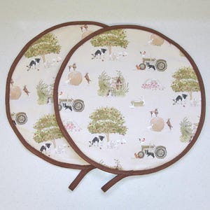 May include: Two round, farm-themed pot holders with a brown border and handle. The cream-colored fabric features a repeating pattern of farm animals, trees, and a tractor. The pot holders are designed to protect surfaces from heat.
