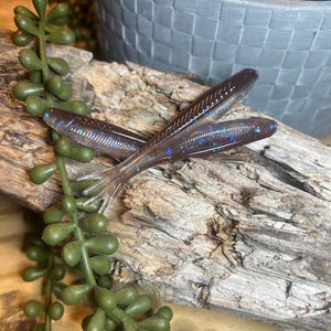 May include: Three soft plastic fishing lures in a dark brown color with blue and silver glitter accents. The lures are lying on a piece of driftwood, with green plant stems and a gray decorative pot in the background.