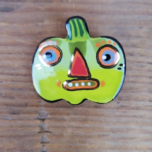 May include: A whimsical, green pumpkin-shaped decorative item with a painted face. The pumpkin features large blue eyes, an orange nose, and a smiling mouth. The design includes orange and gold accents, perfect for Halloween.