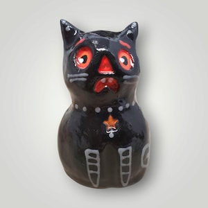 May include: A black ceramic cat figurine with a glossy finish. The cat features bright orange eyes, a matching orange nose, and a red mouth. It has a white dotted collar with an orange star pendant and white skeletal leg markings on its body.