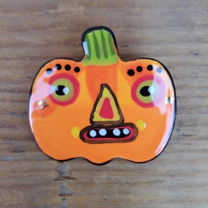 May include: A ceramic pumpkin-shaped pin with a glossy finish. The orange pumpkin has a green stem and a stylized face with black dots, yellow eyes, and a red triangle nose. The pin is outlined in black.
