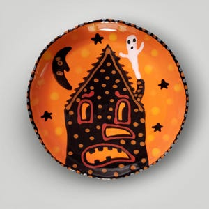 May include: A round ceramic plate featuring a black house with polka dots, orange windows, and a wide orange mouth. The background is a vibrant orange with yellow spots, a black crescent moon, black stars, and a white ghost rising from the chimney.