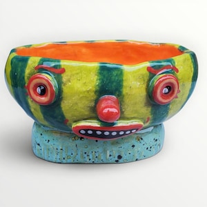 May include: A ceramic bowl featuring a face with a bright orange interior and a green and yellow striped exterior. The face has two round eyes with red rims, a red nose, and a red mouth with white teeth. The base is light blue with black speckles.