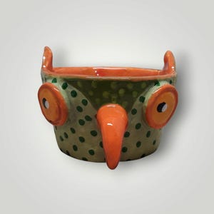 May include: A ceramic bowl shaped like an owl with a light green body featuring dark green polka dots. The bowl has two large orange circular eyes with black pupils, a prominent orange beak protruding from the center, and two small ear tufts.