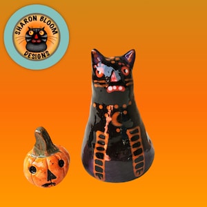 May include: A ceramic Halloween-themed set featuring a black cat figurine with orange accents and a jack-o'-lantern. The cat has a stern expression, and the pumpkin has a carved face. The background is a gradient of orange.