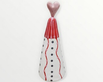 May include: A whimsical, ceramic decorative piece. It features a white, cone-shaped body adorned with black dots and red, wavy lines. The top is red with vertical stripes, and it's topped with a pink heart. The piece has a glossy finish.