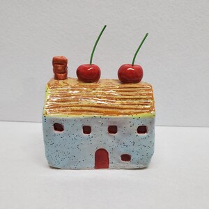 May include: A whimsical ceramic house with a light blue speckled exterior, a red door, and small rectangular windows. The roof is tan with brown lines, and two red cherries with green stems sit on top. A small chimney is on the roof.