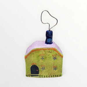 May include: A whimsical ceramic house-shaped photo holder. The house features a green body, a lavender roof, and a dark blue door and chimney. The windows are outlined in yellow. A silver wire extends from the chimney, designed to hold a photo or note.