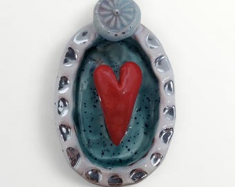 May include: A ceramic heart-shaped trinket dish. The dish features a red heart in the center, surrounded by a teal interior and a light gray border with silver accents. A small, light blue, flower-shaped element is at the top.