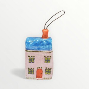 May include: A ceramic house ornament with a blue roof, pink walls, and green windows. The house has an orange door and chimney. A black string is attached for hanging. The ornament is a miniature decorative item.