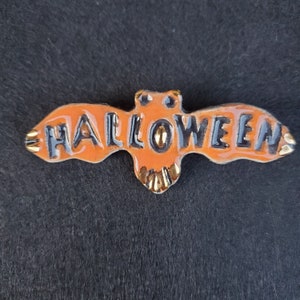 May include: Orange ceramic bat with black lettering that reads "HALLOWEEN". The bat has gold accents.