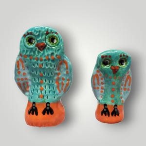 May include: Two ceramic owl figurines. The owls are teal with orange accents and black feet. The owls have large eyes and a small beak. The owls are different sizes, with one being larger than the other.