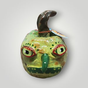 May include: A round, hand-painted ceramic sculpture featuring a light green base with yellow and brown mottled spots. It has a prominent dark green nose, a wide mouth, and two large eyes with red and yellow accents. A dark brown stem protrudes from the top.