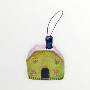 May include: A whimsical, hand-painted ceramic house ornament. The house features a green facade with four sets of windows, a black door, and a lavender roof. A blue chimney and a silver wire loop complete the design.