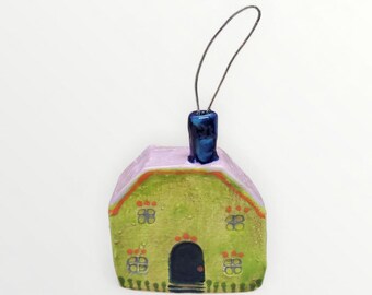 May include: A whimsical, hand-painted ceramic house ornament. The house features a green facade with four sets of windows, a black door, and a lavender roof. A blue chimney and a silver wire loop complete the design.