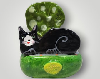 May include: A whimsical ceramic cat figurine with a black body, white face, and green eyes. The cat rests on a green base with a yellow accent. A green, leaf-shaped element is behind the cat.