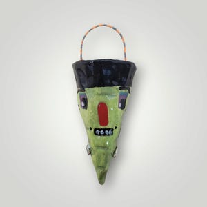 May include: A ceramic wall pocket planter shaped like a green face with a black hair top, a bright red nose, and a small mouth with white teeth. It features a thin, orange and grey striped wire handle attached to the top for hanging.