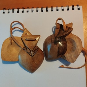 Puede incluir: Se muestran dos instrumentos de percusión de madera, probablemente castañuelas. Cada instrumento presenta un cuerpo en forma de corazón, una parte superior triangular y una caja de resonancia en forma de hoja. La madera tiene un acabado natural, con detalles tallados y correas de cuero.