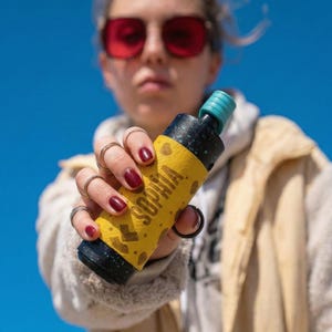 May include: A black and yellow bottle with the name "SOPHIA" printed on it. The bottle has a teal cap and is held against a blue sky background. The bottle is decorated with a yellow wrap with a heart pattern.