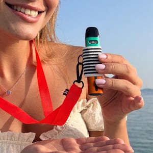 Puede incluir: Una funda para vape de rayas blancas y negras con un vape verde y negro en su interior. La funda está unida a un cordón rojo.