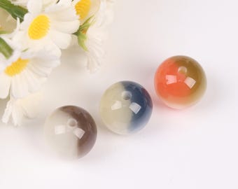 Könnte beinhalten: Drei runde, farbige Perlen mit einem Loch in der Mitte. Eine Perle ist braun und weiß, eine andere weiß und blau, und die dritte orange und gold. Weiße Blumen sind im Hintergrund zu sehen.