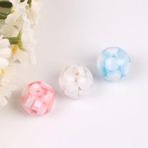 May include: Three round beads in a row. One is pink and white, one is white, and the last is light blue and white. The beads have a hole through the center. The beads are on a white background.