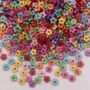 May include: A pile of flower-shaped beads in various colours, each with a black letter or number. The beads are in shades of red, pink, yellow, green, and blue. These small beads are suitable for jewellery making and crafts.