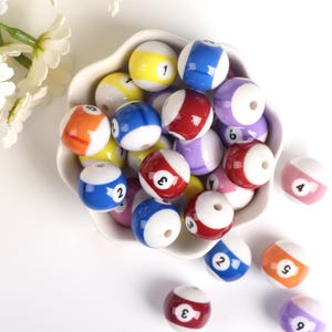 Op de afbeelding: Een kom gevuld met kleurrijke keramische kralen in de vorm van biljartballen, elk met een ander nummer erop gedrukt. De kralen zijn in verschillende kleuren, waaronder rood, blauw, geel, oranje en paars.