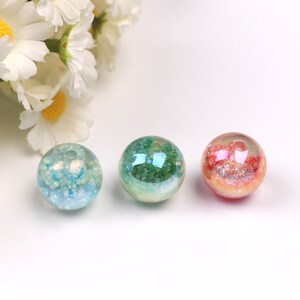 May include: Three round glass beads with a hole in the center. The beads are filled with glitter and have a translucent appearance. The beads are in shades of blue, green, and pink.