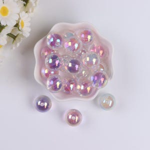 Puede incluir: Un bol lleno de cuentas de vidrio iridiscente en tonos de rosa y morado. Las cuentas son redondas y tienen un acabado liso y brillante.