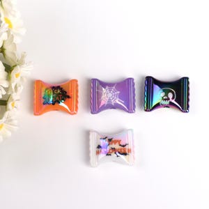 Può includere: Quattro decorazioni di Halloween a forma di caramella iridescente con diversi design. Le decorazioni sono viola, arancioni, bianche e nere. I design includono una ragnatela, "Trick or Treat", pipistrelli e un teschio.