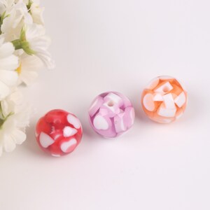 May include: Three round beads with white heart and square shapes embedded in clear resin. The beads are pink, red, and orange.