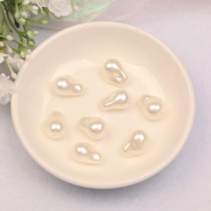 Pode incluir: Uma coleção de pérolas falsas de cor creme, de formato irregular, exibidas em uma tigela rasa de cor creme. As pérolas têm um brilho perolado e lustroso, sugerindo um tema de joias ou materiais de artesanato.
