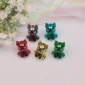 Puede incluir: Seis pequeñas figuras de gato metálicas en varios colores: rojo, plateado, dorado, verde y turquesa. Cada figura tiene un acabado brillante y un diseño de gato estilizado. Las figuras están dispuestas sobre una superficie blanca.