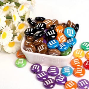 May include: A collection of colorful, candy-shaped beads in a white bowl. The beads are in various colors, including brown, black, orange, blue, green, purple, and yellow. Each bead has a white "m" printed on it. Some beads are scattered around the bowl.