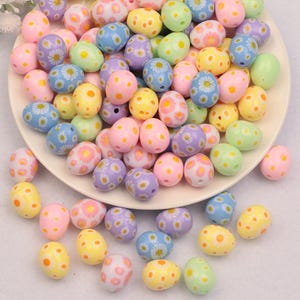 May include: A collection of colorful Easter egg-shaped beads in pastel shades of pink, yellow, blue, green, and purple. The beads are decorated with daisy and polka dot patterns. Some beads are on a white plate.