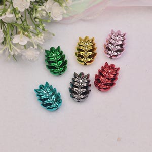 May include: An assortment of metallic leaf-shaped beads in various colors, including green, gold, pink, red, teal, and silver. These decorative beads are arranged on a white surface, with white flowers in the background.